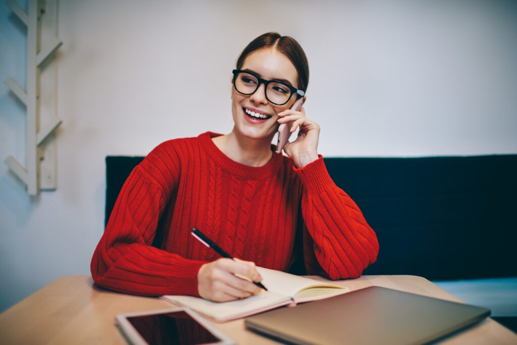 Delighted lady in casual wear and glasses taking notes in notebook while having conversation by smartphone during remote work in cafe