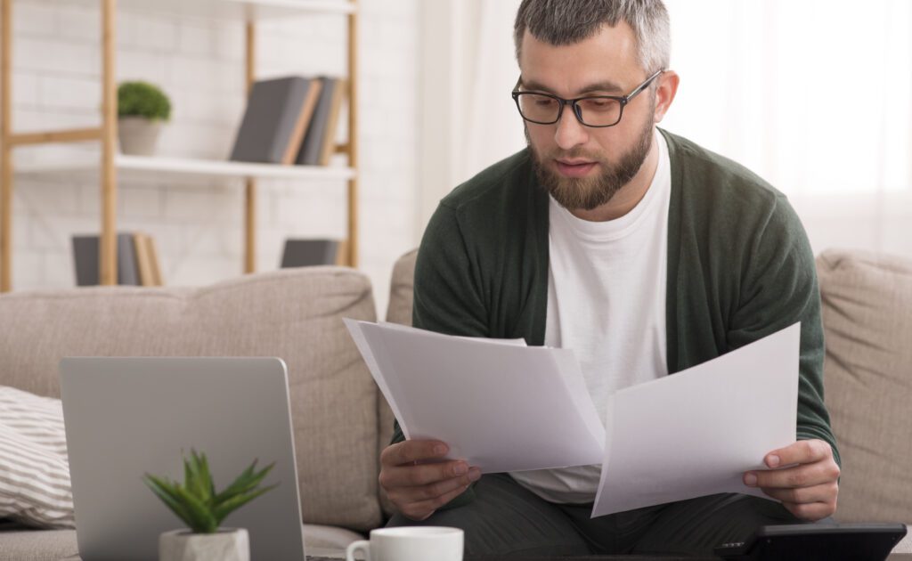 Time to pay the bills. Handsome middle-aged man holding documents and reading it while sitting on the couch at home, copy space